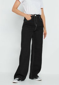 Pantalones de mezclilla de pernera ancha en color negro, con costuras en contraste, cintura alta, bolsillos laterales y cierre de botón. Combinados con zapatillas negras.