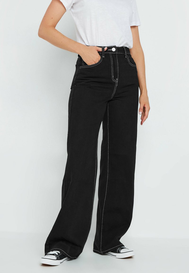 Pantalones de mezclilla de pernera ancha en color negro, con costuras en contraste, cintura alta, bolsillos laterales y cierre de botón. Combinados con zapatillas negras.