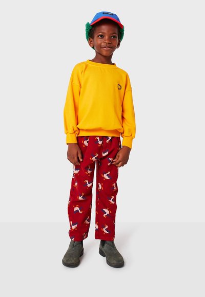 Felpa gialla con un piccolo emblema, pantaloni rossi con motivi di uccelli colorati e stivali grigi; cappelli con accenti blu e rossi.