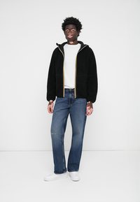 Svart fleecejakke med hvit innerfôr, gul glidelås, sammen med blå jeans og hvite sneakers, stående foran en ensfarget bakgrunn.