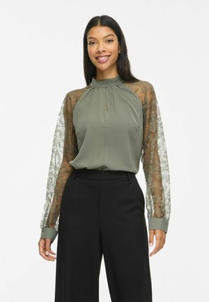 Une femme souriante porte un chemisier vert olive à manches en dentelle transparente et un pantalon noir à jambes larges, sur un fond blanc uni.