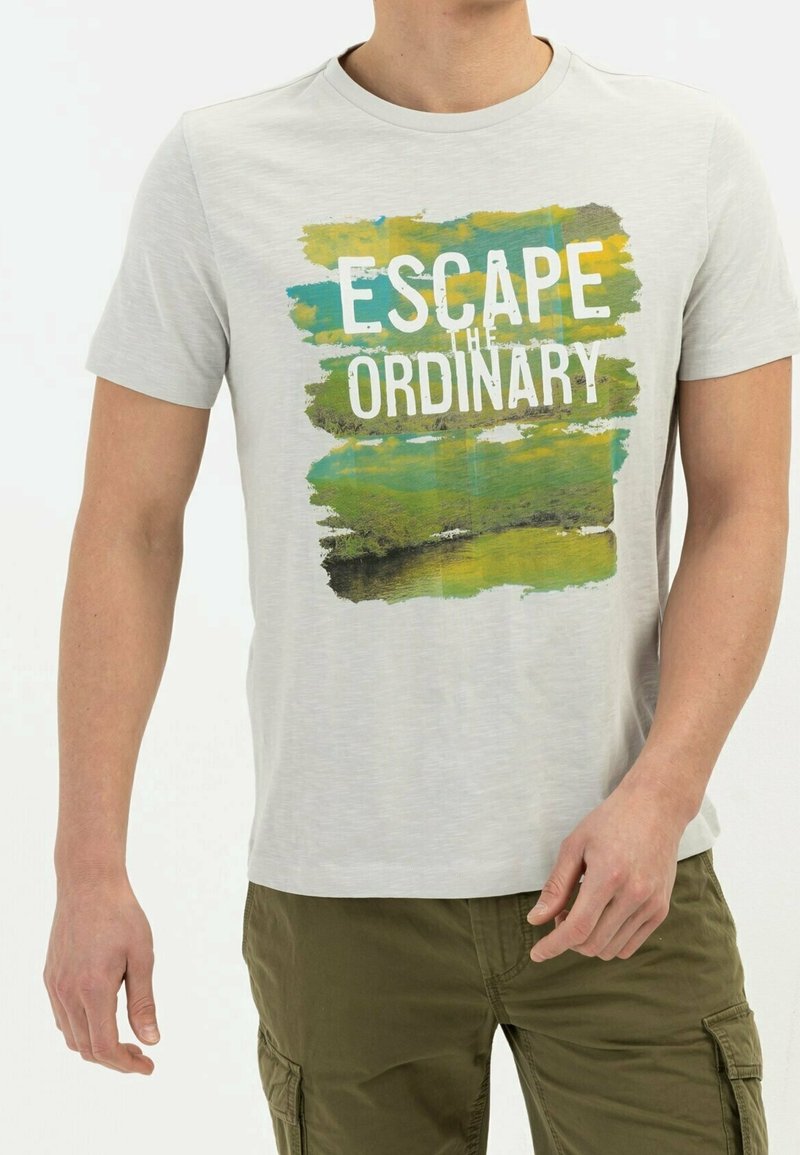 Homme portant un T-shirt gris clair avec un motif nature vert et bleu et le texte « Évadez-vous de l'ordinaire », assorti à un pantalon vert olive.