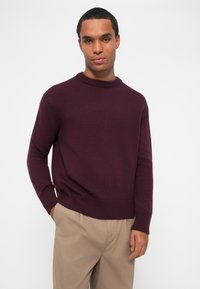 Βουργουνδί πλεκτό πουλόβερ με στρογγυλή λαιμόκοψη, ριμπ μανσέτες και τελείωμα, συνδυασμένο με μπεζ παντελόνι. Λείο ύφασμα, χαλαρή εφαρμογή.
