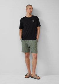 Zwarte t-shirt met logo, lichtgroene shorts en tan sandalen met een bandje. Eenvoudig ontwerp, casual uitstraling en katoenen stof.