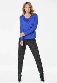 Königsblaues Langarmoberteil mit drapiertem Ausschnitt, kombiniert mit schwarzen gemusterten Hosen und hochhackigen Stiefeletten. Glatte Textur insgesamt.