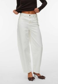 Femme portant un jean blanc taille haute  à jambes larges, un haut à manches longues foncé relevé pour dévoiler le ventre, et des talons pointus marron à pois avec des nœuds.