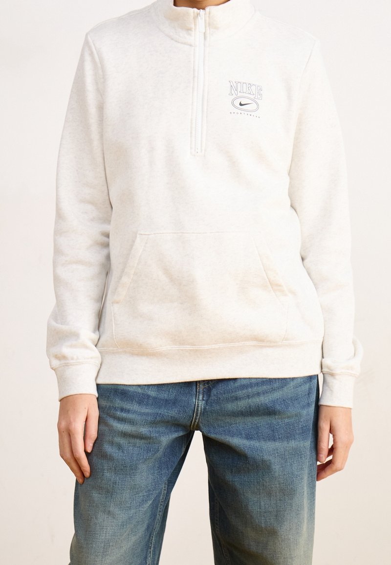 Personne portant un sweat-shirt Nike beige clair avec fermeture éclair à l'avant et poche, associé à un jean en denim bleu, debout devant un fond uni.