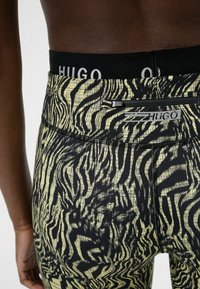 Leggings sportivi neri e gialli con una stampa a zebra testurizzata, dotati di una vita nera con il logo "HUGO" e una tasca con zip sul retro.