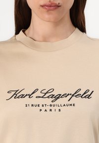 Šviesiai rudos spalvos medvilninis marškinėlis su apvaliu kaklu, su juodu siuvinėtiniu užrašu: "Karl Lagerfeld 21 RUE ST-GUILLAUME PARIS." Lygus paviršius.