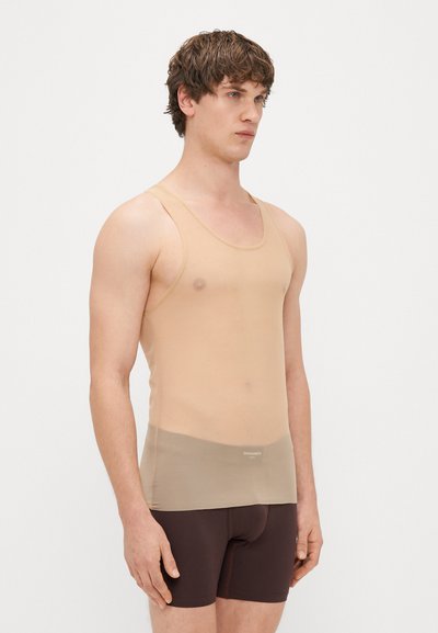 Bēša topiņš no caurspīdīgas auduma, ar izgriezumu ap kaklu, pieguļošu dizainu un smalku logotipa detaļu pie apakšmalas, apvienots ar tumši brūnām šortiem.