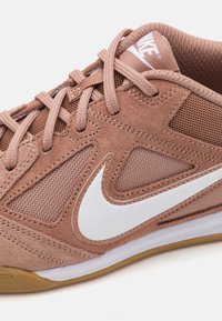 Nike tenisice u svijetlosmeđem semišu i mrežastom materijalu, s bijelim swooshom, okruglim vezicama i gumiranim đonom s teksturiranom površinom.
