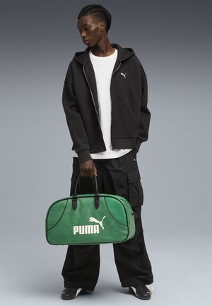 Sac de sport vert Puma avec des accents noirs, affichant un logo blanc bien en vue, une texture lisse et une forme arrondie. Le modèle porte un sweat à capuche noir et un pantalon cargo.