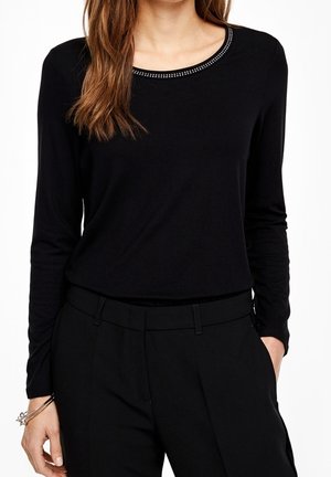 Kvinde iført en sort langærmet skjorte med perlebesat rund halsudskæring og sorte højttaljede bukser, hånd i lommen og armbånd om håndleddet.