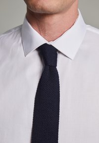Corbata de punto azul marino con una superficie texturizada, usada sobre una camisa blanca con cuello. La corbata es estrecha y presenta un nudo clásico.