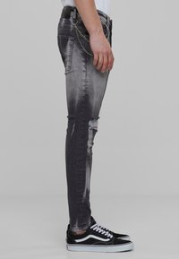 Sieviešu slim-fit džinsi izbalējušā melnā denīmā, ar plēstu dizainu uz ceļa un sudraba ķēdes detaļu sānos. Apvienoti ar melnām sporta kedām.