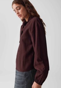 Bordeaux gekleurde blouse van structuurstof met een hoge kraag, doorhangende schouders en lange mouwen. Gepaard met een donkergrijze spijkerrok. Eenvoudig ontwerp, geen patronen.