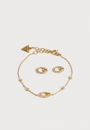 Bracelet Gold Guess avec une fine chaîne, des maillons ovales sertis de cristaux et un fermoir mousqueton, disposé en boucle sur un fond clair.