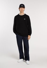 Schwarzes Sweatshirt mit Rundhalsausschnitt und gesticktem Logo, kombiniert mit dunkelblauen Jeans und weißen Sneakers, im lässigen Schnitt.