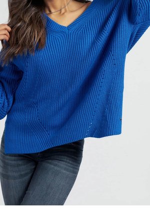 Femme portant un pull bleu texturé à col en V et un jean gris foncé, un bras levé et de longs cheveux ondulés sur l'épaule.