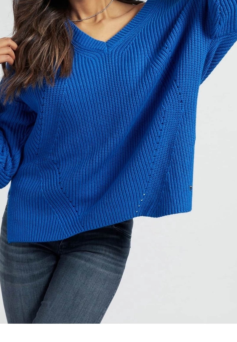 Femme portant un pull bleu texturé à col en V et un jean gris foncé, un bras levé et de longs cheveux ondulés sur l'épaule.