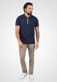 Marineblå poloskjorte med korte ermer og lynlås i halsen, kombinert med beige chinos og hvite joggesko, med en glatt tekstur og enkelt design.