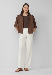 Brown linen short-sleeve shirt over a brown tank top, paired with white, loosely fitted, textured pants and brown sandals.