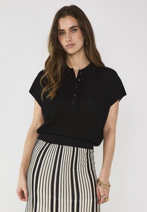 Femme aux longs cheveux bruns portant un haut noir à manches courtes avec col et une jupe taille haute à rayures verticales noires et blanches, accessoirisée de bracelets.