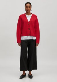 Roter Strickcardigan mit V-Ausschnitt, fünf Holzknöpfen und weiten Ärmeln, kombiniert mit einem weißen Hemd und schwarzen weit geschnittenen Hosen.