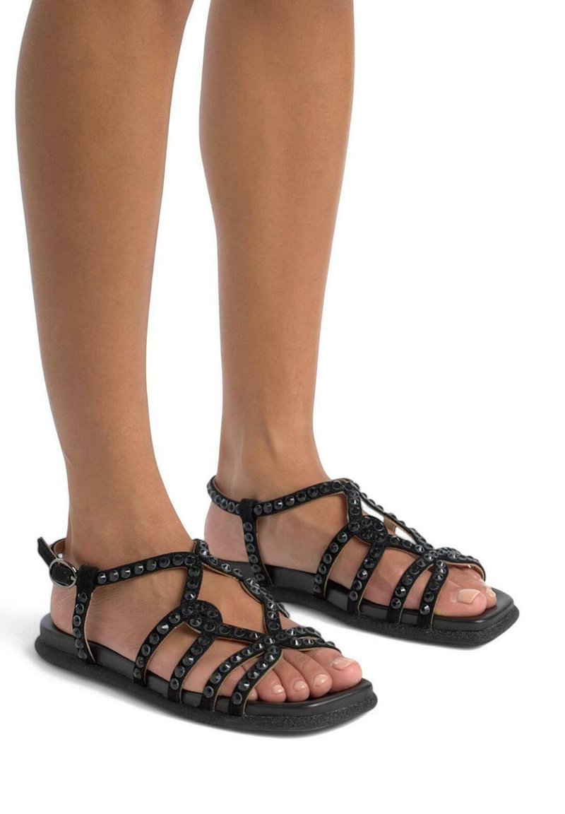 Czarne sandały na płaskiej podeszwie z paskami, charakteryzujące się krzyżującym się wzorem i dekoracyjnymi ćwiekami, zapinane na regulowany pasek w okolicy kostki.