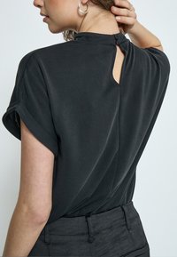 Top nero a maniche corte con colletto alto, chiusura con bottoni sul retro e una piccola apertura. Realizzato in tessuto liscio e leggero.
