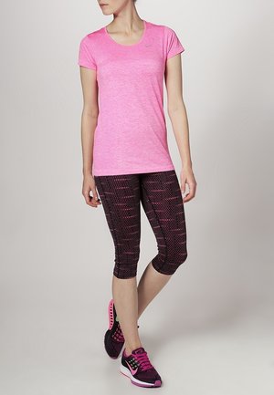 Vrouw draagt een roze sportshirt met korte mouwen, zwart-roze caprileggings met patroon en zwart-roze hardloopschoenen, staande met gekruiste benen.