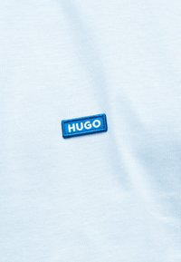 Ανοιχτό μπλε ύφασμα με ένα μικρό ορθογώνιο μπλε κέντημα με λευκά γράμματα "HUGO" κοντά στο κέντρο.