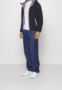 Uomo che indossa una felpa nera con zip, una maglietta bianca, jeans blu larghi e sneakers bianche, in piedi contro uno sfondo semplice.