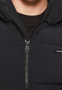 Czarna kurtka puchowa z kapturem, z widocznym zapięciem na zamek błyskawiczny, teksturowanym materiałem oraz naszywką z logo Calvin Klein na piersi.