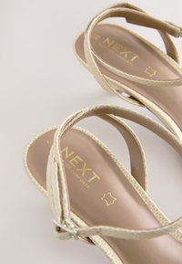 Sandales à brides beiges avec une partie supérieure texturée, une semelle plate et un logo doré sur la semelle intérieure. Bride réglable à la cheville avec une boucle en métal argenté.