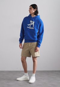 Felpa blu con logo, abbinata a pantaloni corti color khaki e sneakers bianche. Il tessuto appare morbido, con una tasca a marsupio e lacci.