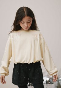 Krémový svetr s flitrovými detaily na ramenou, dlouhými rukávy a nařaseným pasem, doplněný o černé třpytivé šortky.