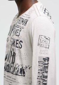 Langarmshirt aus hellbeigem Stoff mit schwarzem Drucktext und Grafiken, verziert mit Distressed-Details und roten Akzentnähten an der Schulter.