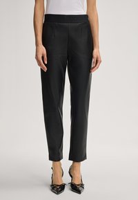 Woman wearing black faux leather tapered pants with a high waist and black pointed-toe heels against a plain background.