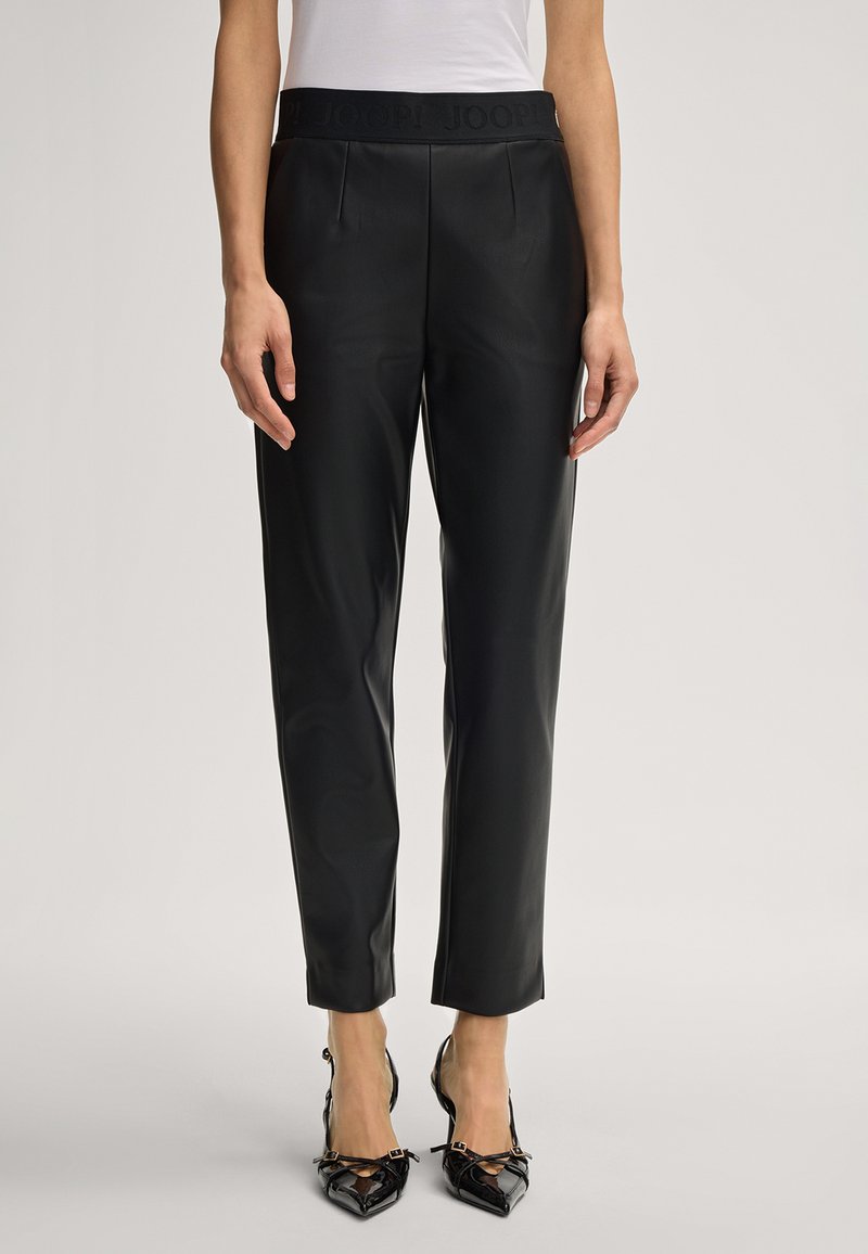 Woman wearing black faux leather tapered pants with a high waist and black pointed-toe heels against a plain background.