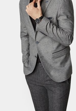 Homme ajustant son col, portant un blazer gris, une chemise noire, un pantalon à carreaux et une montre-bracelet noire avec un bracelet en cuir noir.