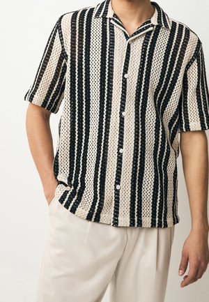Man draagt een beige en zwarte kortärmelig overhemd met verticale strepen, een gebreide textuur en lichtgekleurde broek.