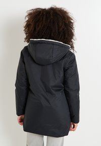 Parka negra acolchada con capucha, con un forro de piel sintética blanca y un corte de longitud media, diseñada para brindar calidez y comodidad.