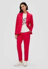 Bright pink suit with a tailored blazer and matching trousers, paired with a white t-shirt featuring a geometric black and red pattern. White sneakers.