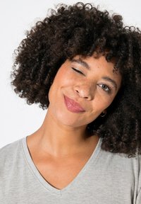Retrato de una persona con cabello rizado y oscuro, y pecas, que lleva una camiseta de cuello en V gris claro. Está guiñando un ojo y sonriendo levemente.