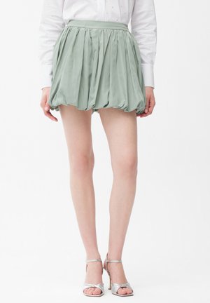 Kobieta w jasnozielonej plisowanej mini spódnicy balonowej, białej bluzce na długi rękaw i srebrnych sandałach na wysokim obcasie.