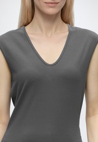 Femme aux longs cheveux blonds porte un haut gris foncé sans manches et à col en V. La photo est recadrée sous le nez et au-dessus de la taille.