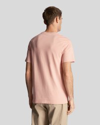 Un homme portant un t-shirt rose clair à manches courtes uni et un pantalon beige, vu de dos sur un fond gris clair.