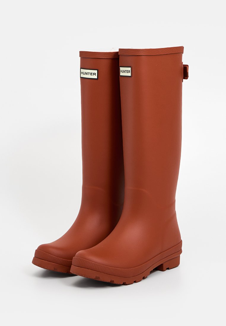 Botas de lluvia de goma Hunter en color óxido, diseño alto, fabricadas con un material suave y duradero, con acentos de logo y suelas texturizadas para un mejor agarre.
