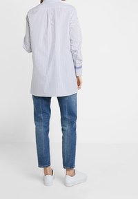 Gestreifte Hemdjacke in Weiß und Marineblau, mit langen Ärmeln, kombiniert mit blauen Jeans und weißen Sneakers. Fokus auf die Rückansicht und einen lässigen Schnitt.